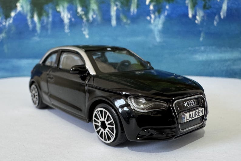 Peut inclure: Une voiture jouet Audi A1 noire avec un toit argent&eacute; et des roues blanches. La voiture porte le nom "LAUREN" sur sa plaque d'immatriculation. L'arri&egrave;re-plan est une image floue bleue et blanche.