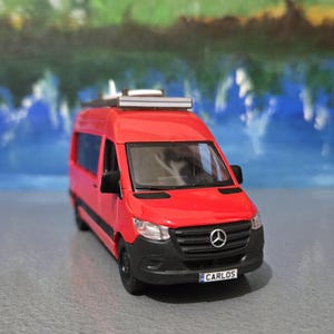 May include: A red toy van with black accents, a roof rack, and the word "CARLOS" on the license plate. The van features a Mercedes-Benz logo on the front and is set against a blurred background.