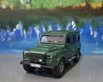 May include: A dark green Land Rover Defender model car with black accents. The model has a license plate that reads "FRED 78". The car is set against a blurred background of blue and green.
