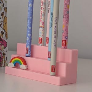 May include: A pink tiered pen holder with six pens and a rainbow figurine. The pens are decorated with various designs and text, including "Believe in Magic" and "You are Beautiful". A hardcover book with colorful illustrations is visible.