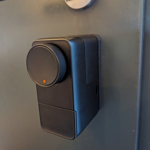May include: A black smart door lock with a round, black knob and a small orange light. The lock is mounted on a gray door.