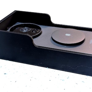 Könnte beinhalten: Eine schwarze, rechteckige Dockingstation mit einer gebogenen Vorderkante. Sie hält ein silbernes Gerät mit einer kreisförmigen Oberseite und einem Kameraobjektiv. Auf dem Gerät steht "AirTag".