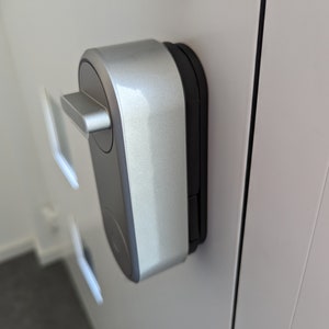 Puede incluir: Una cerradura de puerta inteligente plateada con un teclado negro y una manija plateada. La cerradura está montada en una puerta blanca.