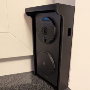 May include: A black smart doorbell system mounted on a wall. The system includes a camera, a circular button with a bell icon, and a protective housing. The doorbell is designed for home security and communication.