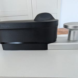 Op de afbeelding: Een zwart Acoara smart home-apparaat met een zilveren knop aan de zijkant. Het apparaat is gemonteerd op een wit oppervlak.