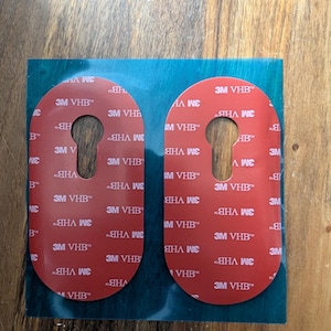 May include: Two red adhesive pads with a keyhole-shaped cutout in the center. Each pad features the white text 3M VHB printed repeatedly across the surface. The pads are mounted on a rectangular teal-colored backing sheet for protection.