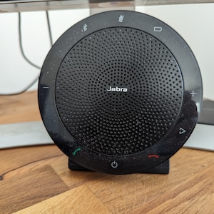 Könnte beinhalten: Ein schwarzes Jabra-Lautsprechertelefon mit Mikrofon und Anrufsteuertasten. Das Lautsprechertelefon befindet sich auf einer Holzoberfläche.
