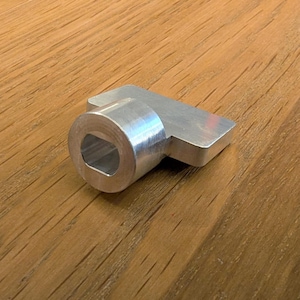 May include: A silver metal component featuring a hexagonal aperture and a rectangular handle. The object, made of a shiny, reflective material, rests on a wooden surface.