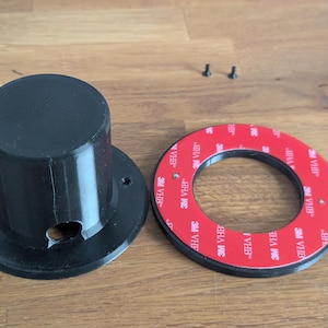 Könnte beinhalten: Ein schwarzes zylindrisches Bauteil mit kreisförmiger Basis, Schraubenlöchern und einer kleinen Öffnung, neben einem schwarzen Ring mit rotem Klebeband. Zwei kleine Schrauben sind ebenfalls sichtbar. Es handelt sich um Teile eines Montagesystems.