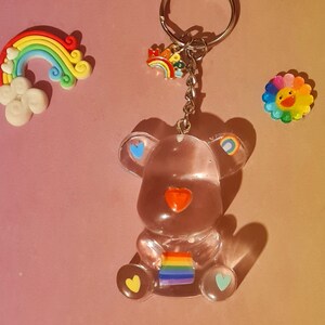 May include: A clear bear-shaped keychain with rainbow accents, holding a rainbow flag. The keychain has a silver ring and chain. Additional rainbow and flower-shaped charms are also present. A rainbow and cloud decoration is also visible.