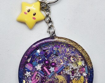 Può includere: Un portachiavi a tema celeste con un ciondolo a forma di stella gialla e un pendente rotondo pieno di glitter, stelle e perline. Il pendente ha un gradiente di viola, blu e oro. Il portachiavi è attaccato a un anello d'argento.