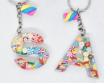 May include: Two keychains with the letters "S" and "A". The letters are filled with colorful candies and sprinkles, and attached to a silver keyring. Each keychain has a small, colorful, swirled candy charm.