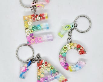 May include: Three resin keychains spelling out "EAC". Each letter is filled with colorful fruit slices and attached to a silver keyring. The keychains also feature small, decorative charms.