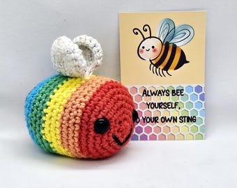 May include: A handmade, crocheted bee in rainbow colors with a smiling face and white wings. Beside it is a card with a cartoon bee illustration and the text "ALWAYS BEE YOURSELF, YOUR OWN STING."