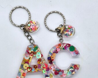 May include: Two keychains with clear resin letters A and C filled with colorful glitter and sequins. Each keychain has a small clear glass orb filled with colorful glitter attached to the top.