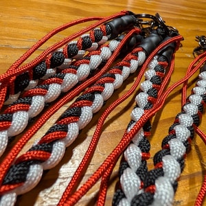 May include: Several lanyards crafted from braided paracord in a repeating pattern of red, black, and white. Each lanyard features a black rubber connector at the top, attached to a circular metal ring, with loose red cord strands extending outwards.