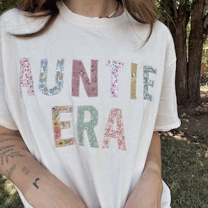 Könnte beinhalten: Ein weißes T-Shirt mit den Worten "Auntie Era", die in bunten Stoff-Patches ausgeschnitten sind. Die Patches bestehen aus Blumenmustern in den Farben Rosa, Blau, Grün und Gelb.