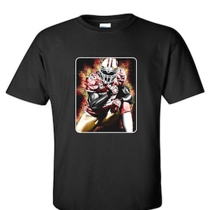 May include: Black t-shirt featuring a graphic of a football player in a red and gold uniform, kneeling in a dynamic pose. The player is wearing a helmet and pads, set against a fiery, colorful background. The design is enclosed in a white rectangle.