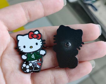 May include: Two enamel pins featuring a cartoon cat character. One pin shows the character wearing a green soccer jersey with the number 4 and a red bow, standing next to a soccer ball. The other pin shows the solid black back of the same character.