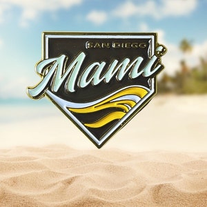 May include: Gold baseball-shaped pin with a black background and white and yellow waves. The text "San Diego" and "Mami" are written in white on the pin.