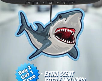 May include: A shark-shaped car air freshener hanging from a rearview mirror. The shark is grey and white with an open mouth showing sharp teeth. A blue starburst graphic includes the text Buy 2 Get 1 Free and Extra Scent Bottle Included.