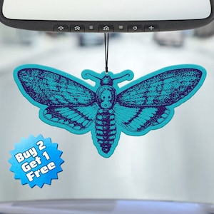 May include: A teal blue car air freshener shaped like a moth with a skull design. The air freshener is hanging from a rearview mirror. The text "Buy 2 Get 1 Free" is on a blue starburst.