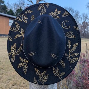 May include: A black fedora hat with gold leaf and crescent moon designs. The hat features a wide brim and a structured crown. The gold leaf pattern is painted around the brim and on the crown. A stylish accessory for any outfit.