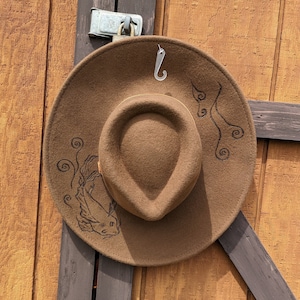 May include: A brown felt fedora hat with a wide brim and a black embroidered design of a fish and swirling lines. The hat is hanging on a hook.