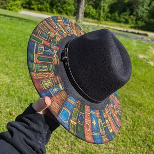 Op de afbeelding: Zwarte fedora hoed met een rand versierd met een kleurrijk ontwerp van gestapelde boeken. De hoed heeft een zwarte leren band en een gesp. De rand is versierd met boekruggen in blauw, bruin en goud.