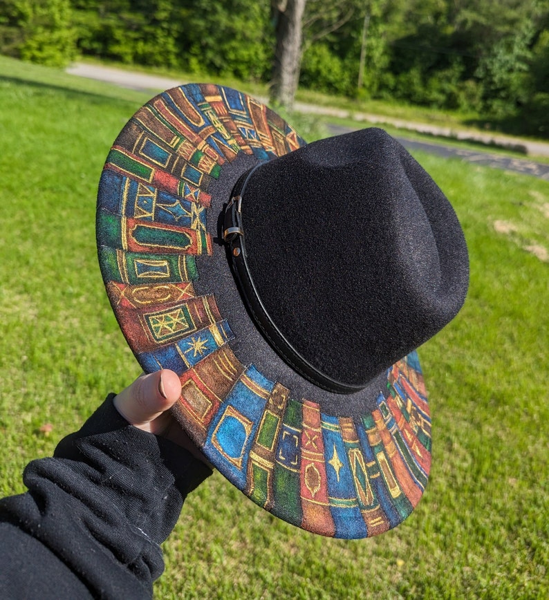 Op de afbeelding: Zwarte fedora hoed met een rand versierd met een kleurrijk ontwerp van gestapelde boeken. De hoed heeft een zwarte leren band en een gesp. De rand is versierd met boekruggen in blauw, bruin en goud.