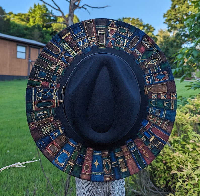 Op de afbeelding: Een zwarte fedora hoed met een decoratieve rand met een kleurrijk patroon van boeken in verschillende vormen en maten. De boeken zijn in tinten blauw, rood en bruin, met gouden accenten. De hoed wordt buiten gepresenteerd.