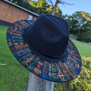 Op de afbeelding: Een zwarte fedora hoed met een patroonrand. De rand heeft een kleurrijk geometrisch ontwerp met blauwe, rode en gouden accenten. De hoed wordt buiten op een houten paal gepresenteerd.