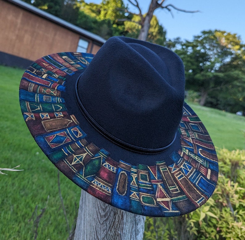 Op de afbeelding: Een zwarte fedora hoed met een patroonrand. De rand heeft een kleurrijk geometrisch ontwerp met blauwe, rode en gouden accenten. De hoed wordt buiten op een houten paal gepresenteerd.