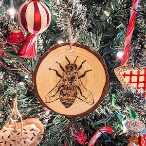 May include: A wooden Christmas ornament with a burnt image of a bee on it. The ornament is hanging on a Christmas tree.