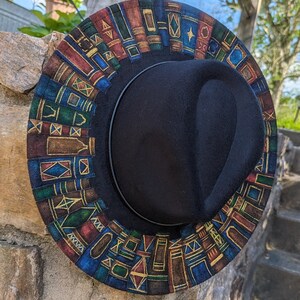 Op de afbeelding: Een zwarte fedora hoed met een rand versierd met een kleurrijk patroon van boeken. De boeken zijn in tinten blauw, groen, rood en bruin, met gouden accenten. De hoed rust op een stenen muur.