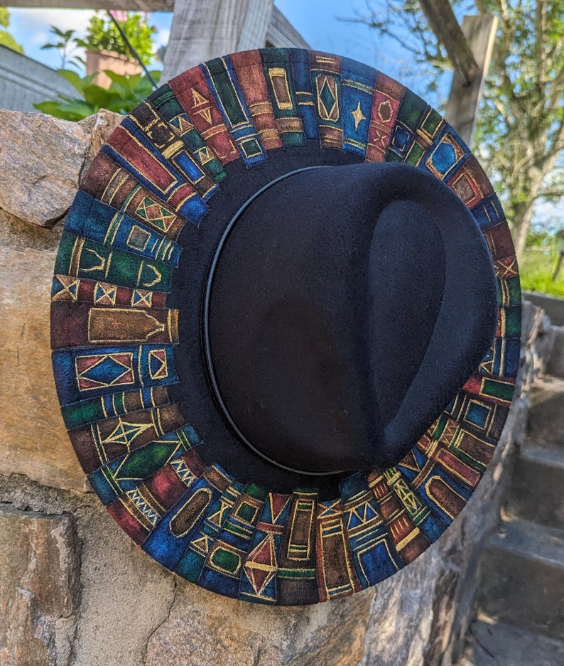 Op de afbeelding: Een zwarte fedora hoed met een rand versierd met een kleurrijk patroon van boeken. De boeken zijn in tinten blauw, groen, rood en bruin, met gouden accenten. De hoed rust op een stenen muur.