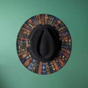 Op de afbeelding: Een zwarte fedora hoed met een kleurrijke rand met een herhalend patroon van gestileerde boeken in tinten blauw, rood en goud. De hoed hangt tegen een effen groene achtergrond.