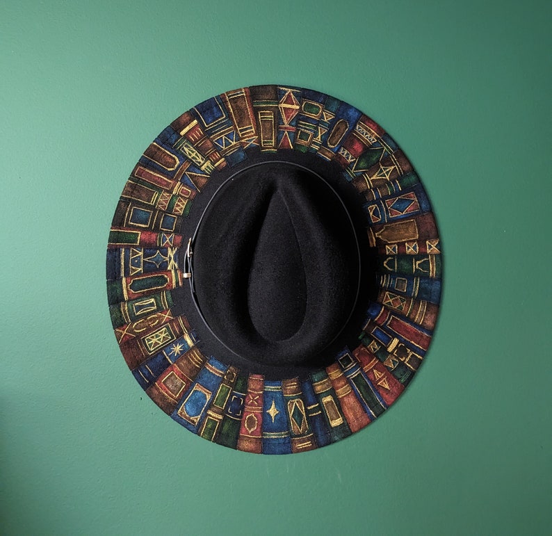 Op de afbeelding: Een zwarte fedora hoed met een kleurrijke rand met een herhalend patroon van gestileerde boeken in tinten blauw, rood en goud. De hoed hangt tegen een effen groene achtergrond.