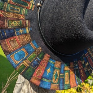Op de afbeelding: Zwarte fedora hoed met een decoratieve rand met een kleurrijk, handgeschilderd ontwerp van gestapelde boeken. De boeken zijn in tinten blauw, groen, rood en bruin met gouden accenten. Een zwarte leren band met een gouden gesp omcirkelt de hoed.