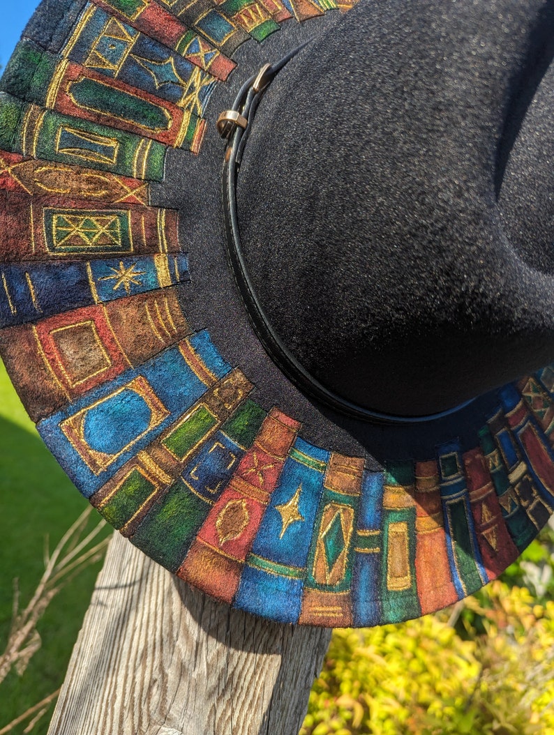 Op de afbeelding: Zwarte fedora hoed met een decoratieve rand met een kleurrijk, handgeschilderd ontwerp van gestapelde boeken. De boeken zijn in tinten blauw, groen, rood en bruin met gouden accenten. Een zwarte leren band met een gouden gesp omcirkelt de hoed.