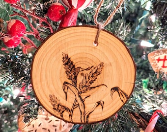May include: A wood slice Christmas ornament with a burnt design of wheat stalks. The ornament is hanging on a Christmas tree.