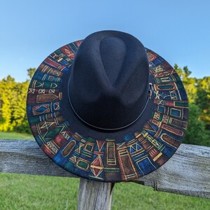 Op de afbeelding: Een zwarte fedora hoed met een patroonrand met geometrische vormen in blauw, rood, goud en groen. De hoed rust op een houten hek, met een achtergrond van groene bomen en een blauwe lucht.