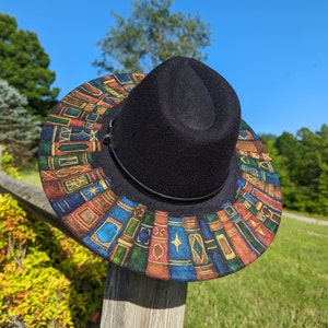 Op de afbeelding: Een zwarte fedora hoed met een rand versierd met een kleurrijk patroon van boeken. De hoed rust op een houten hekpaal tegen een achtergrond van groene bomen en een heldere blauwe lucht.