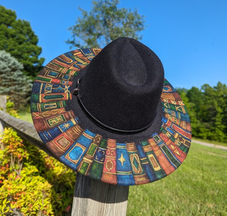 Op de afbeelding: Een zwarte fedora hoed met een rand versierd met een kleurrijk patroon van boeken. De hoed rust op een houten hekpaal tegen een achtergrond van groene bomen en een heldere blauwe lucht.