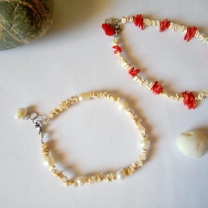 Può includere: Due braccialetti con perline naturali. Uno presenta dettagli in corallo rosso, l'altro perle. Entrambi sono su una superficie bianca, con una piccola pietra bianca e una roccia verde e grigia sullo sfondo.