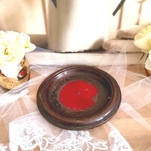 Puede incluir: Un pequeño plato redondo de cerámica marrón con un centro rojo vibrante. El plato está sobre un paño de encaje blanco, con un fondo borroso con flores y objetos decorativos. El plato probablemente se utiliza con fines decorativos.