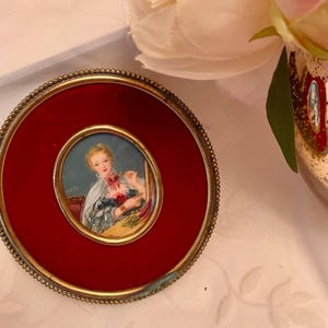 May include: A miniature portrait in a gold-coloured frame. The portrait depicts a woman in a blue and white dress with a red bow. The frame has a red velvet background and a beaded edge.