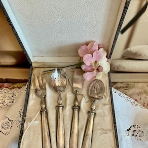May include: A set of vintage silver cutlery, comprising a fork, slotted spoon, knife, and serving spoon, displayed in a decorative box. The silver utensils feature ornate handles and are arranged on cream fabric. Pink and white flowers provide a colourful accent.
