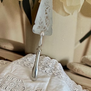 Puede incluir: Un servidor de pastel plateado con un mango adornado y un diseño decorativo calado en la hoja. La hoja presenta patrones florales y de volutas. El servidor descansa sobre un paño blanco con ribete de encaje.