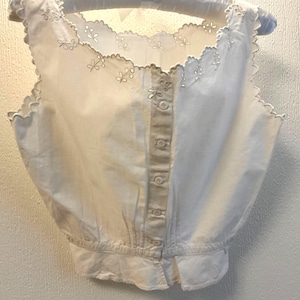 May include: A white sleeveless blouse with a scalloped neckline and floral embroidery. The blouse has a button-down front and a gathered waist. It is hanging on a white padded hanger.